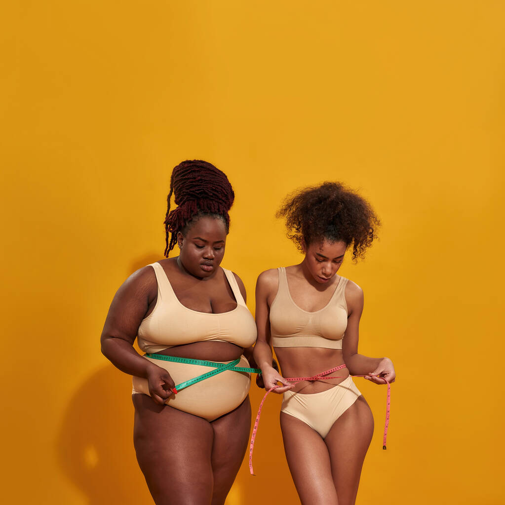 Studio záběr dvou afrických Američanek s různou tělesnou hmotností měřící jejich pas pomocí metrické pásky opatření, stojící spolu izolované nad oranžovým pozadím - Fotografie, Obrázek