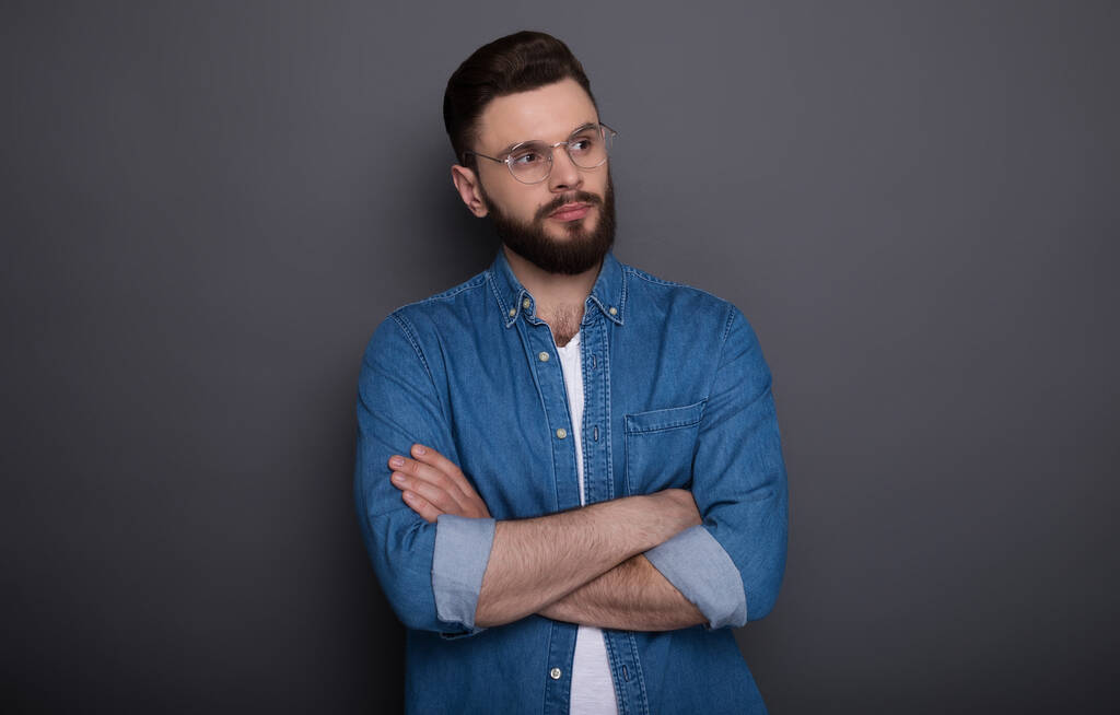 Уверенный умный современный бородатый мужчина в джинсовой одежде и очках позирует на сером фоне в студии - Фото, изображение