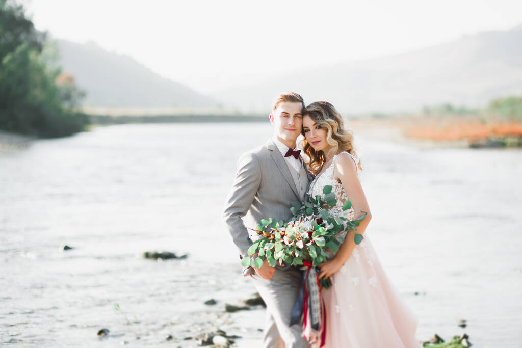 Hermosa pareja de boda besándose y abrazándose cerca de la orilla de un río de montaña con piedras - Foto, Imagen