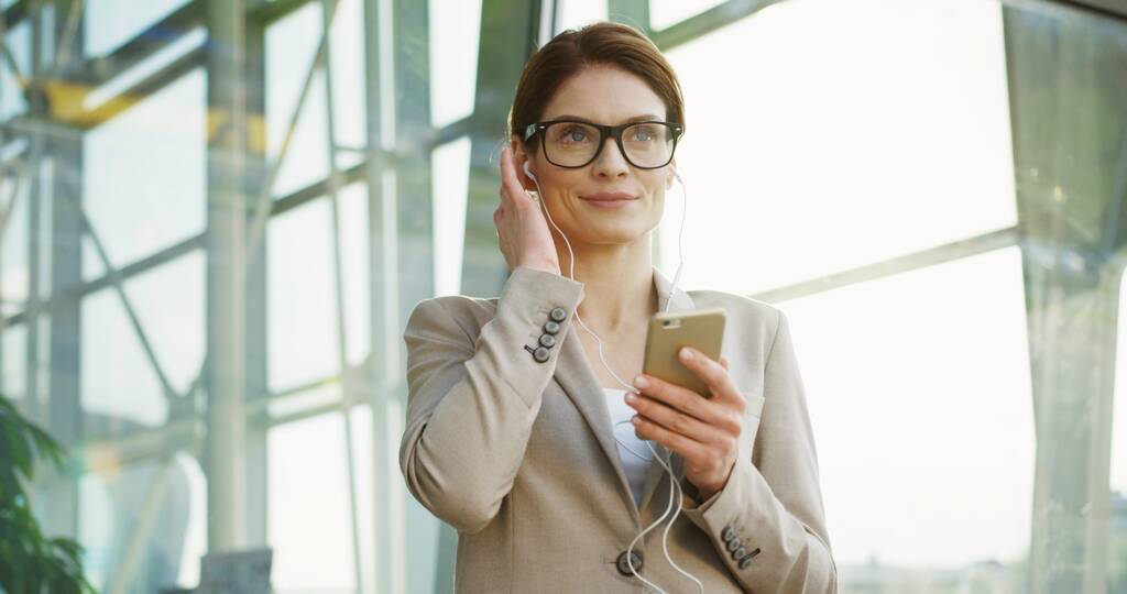 Portrétní záběr atraktivní bělošky v brýlích, jak si nasazuje sluchátka, nahrává smartphone a poslouchá hudbu na pozadí skleněné budovy. Venku - Fotografie, Obrázek