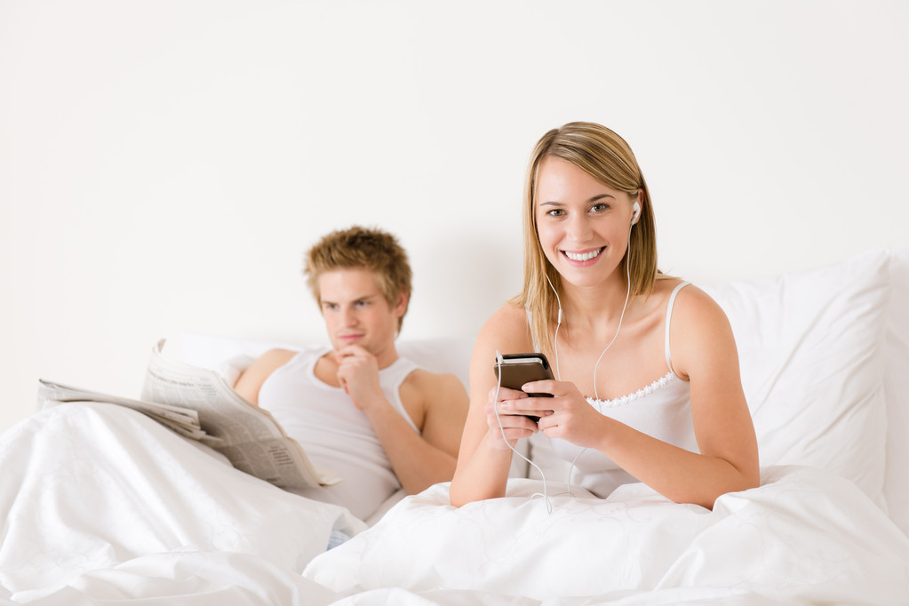 νεαρό ζευγάρι χαλαρώσουν στο λευκό κρεβάτι να ακούσετε μουσική, να διαβάσει εφημερίδα - Φωτογραφία, εικόνα