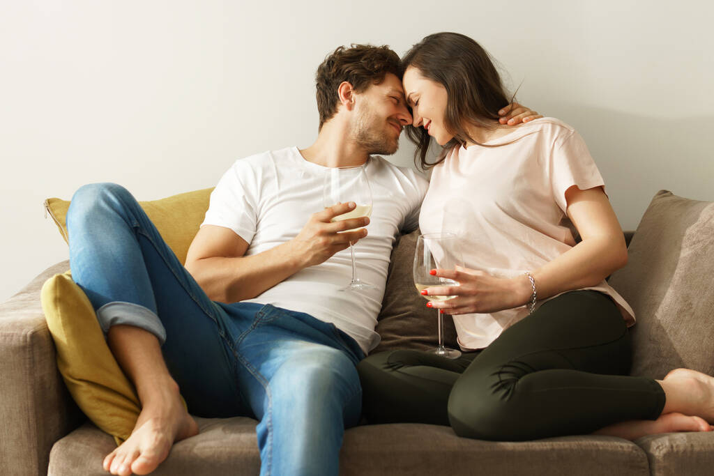 Νεαρό και ευτυχισμένο ζευγάρι πίνοντας κρασί και χαλαρώνοντας στο σπίτι - Φωτογραφία, εικόνα
