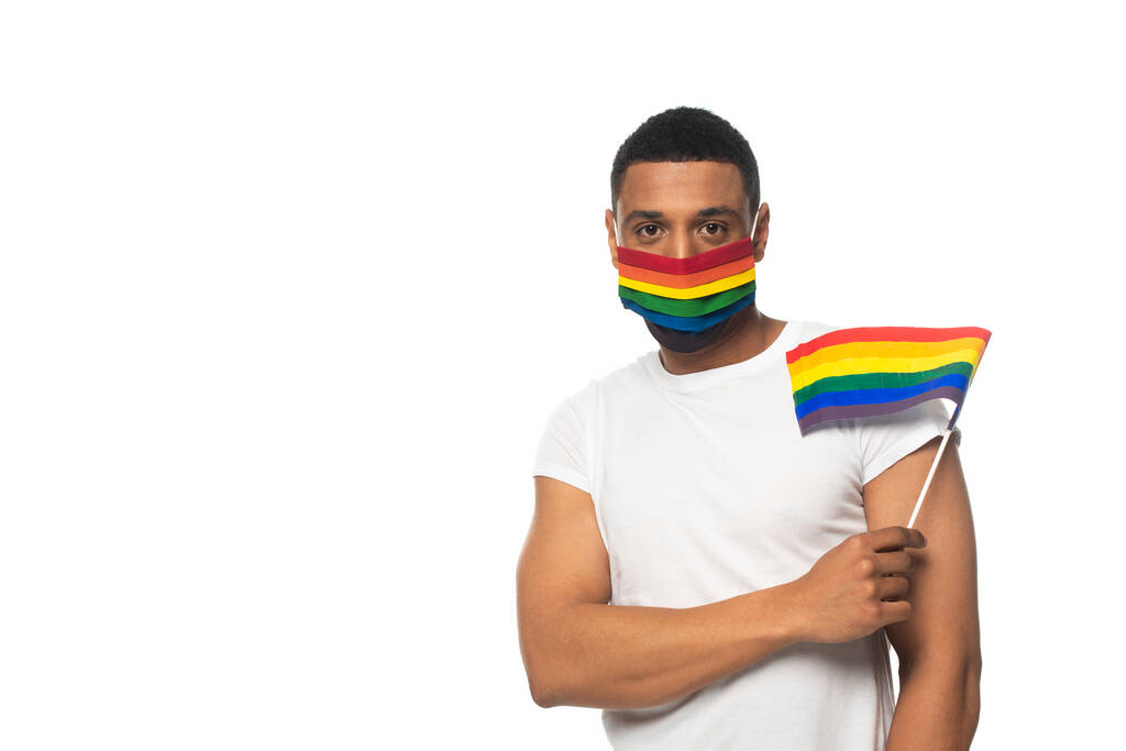 Afrikanisch-amerikanischer Mann trägt Regenbogenfarben medizinische Maske und hält kleine lgbt Flagge isoliert auf weiß - Foto, Bild