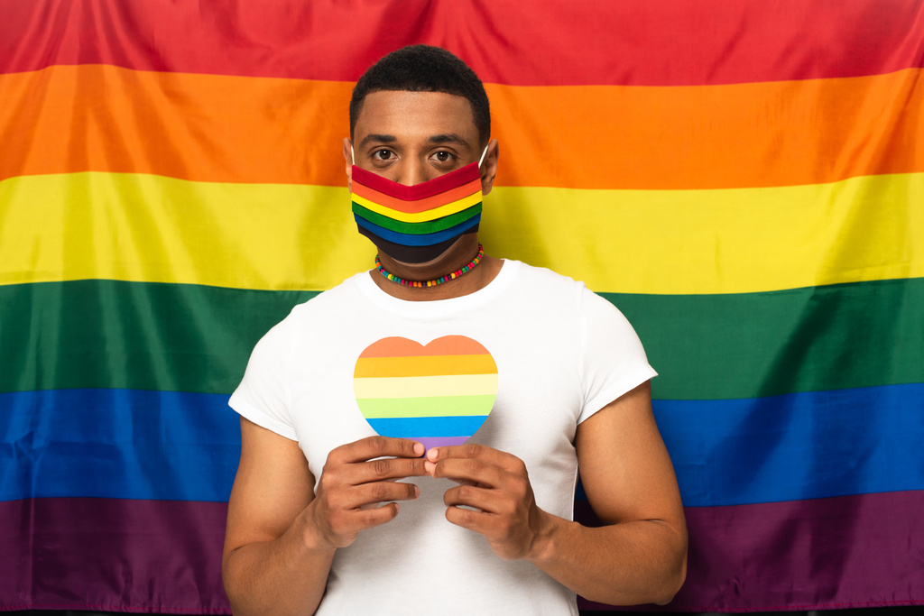 homem americano africano com máscara de segurança e coração de papel em cores do arco-íris no fundo da bandeira lgbt - Foto, Imagem