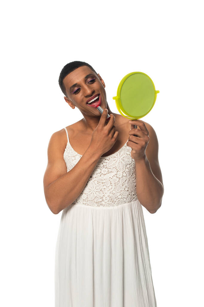 Afričanky americká transgender žena v slunečních šatech nanášení rtěnky při pohledu do zrcadla izolované na bílém - Fotografie, Obrázek