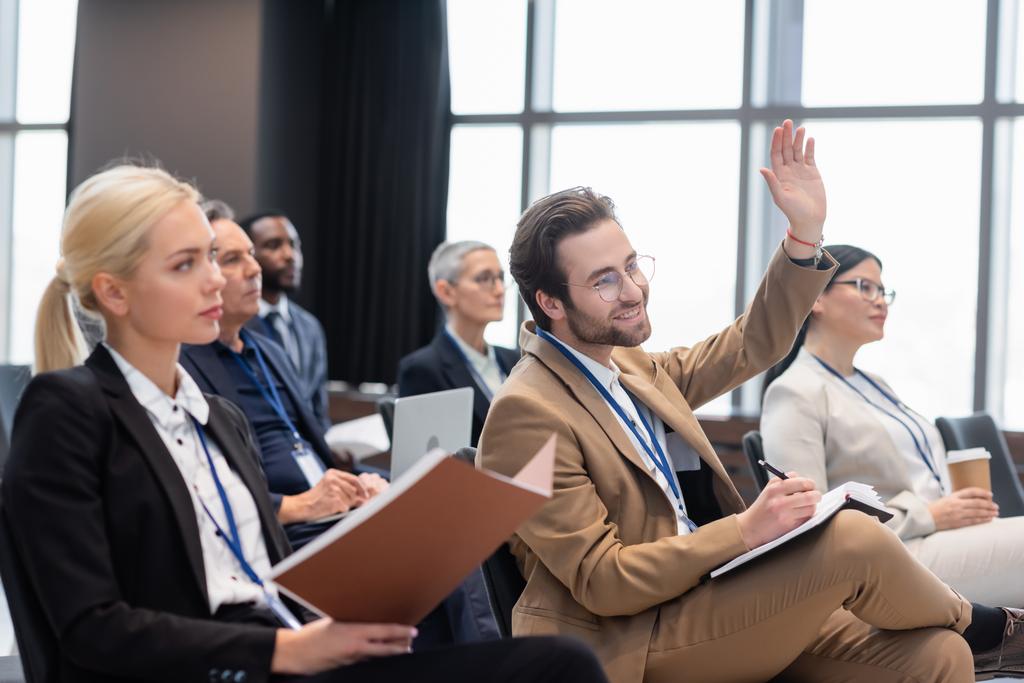Улыбающийся бизнесмен поднимает руку во время семинара рядом с межрасовыми коллегами  - Фото, изображение