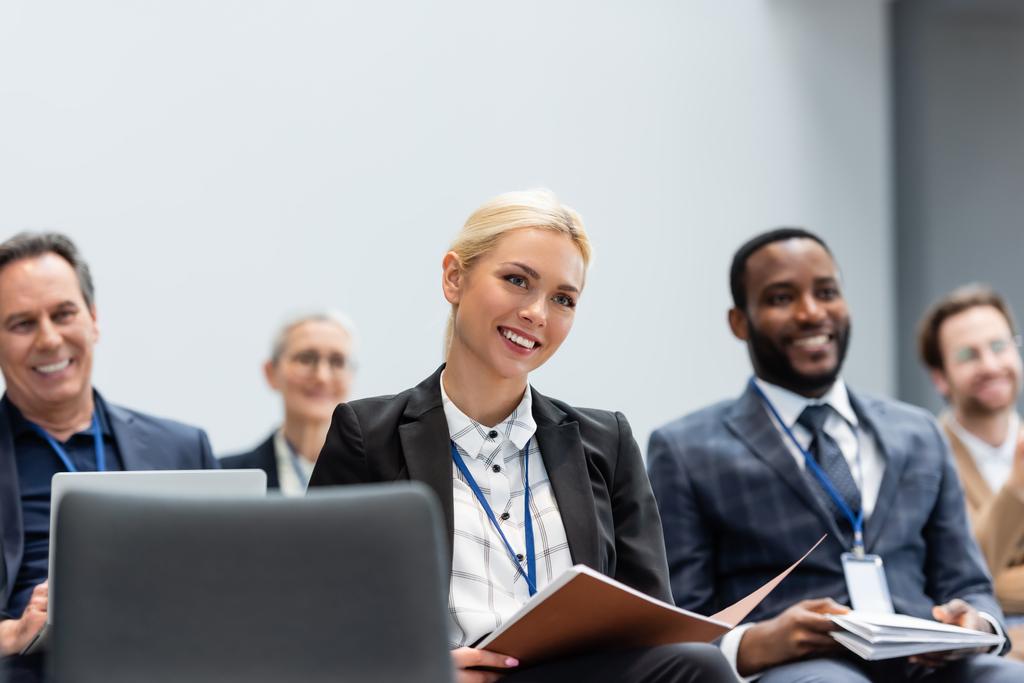Веселая деловая женщина с бумажной папкой сидит рядом с многонациональными коллегами во время семинара  - Фото, изображение