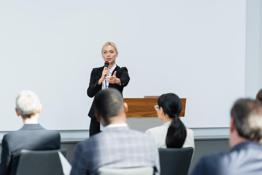 young lecturer talking in microphone while pointing with finger at business people on blurred foreground - Photo, Image