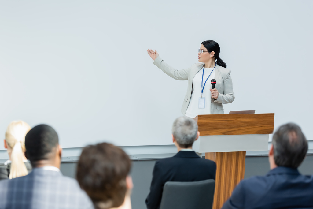 Asijský lektor drží mikrofon a ukazuje rukou v blízkosti rozmazané publikum během semináře - Fotografie, Obrázek