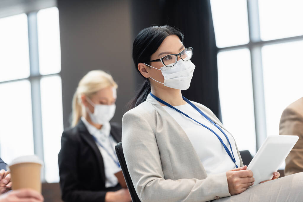 Ασιάτισσα επιχειρηματίας με ιατρική μάσκα κρατώντας ψηφιακή ταμπλέτα κατά τη διάρκεια σεμιναρίου - Φωτογραφία, εικόνα