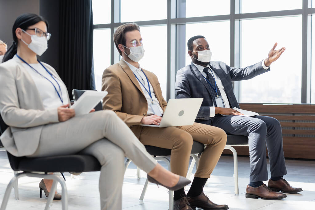 Afrikaans Amerikaans zakenman met medisch masker wijzend met de hand in de buurt van collega 's met gadgets - Foto, afbeelding