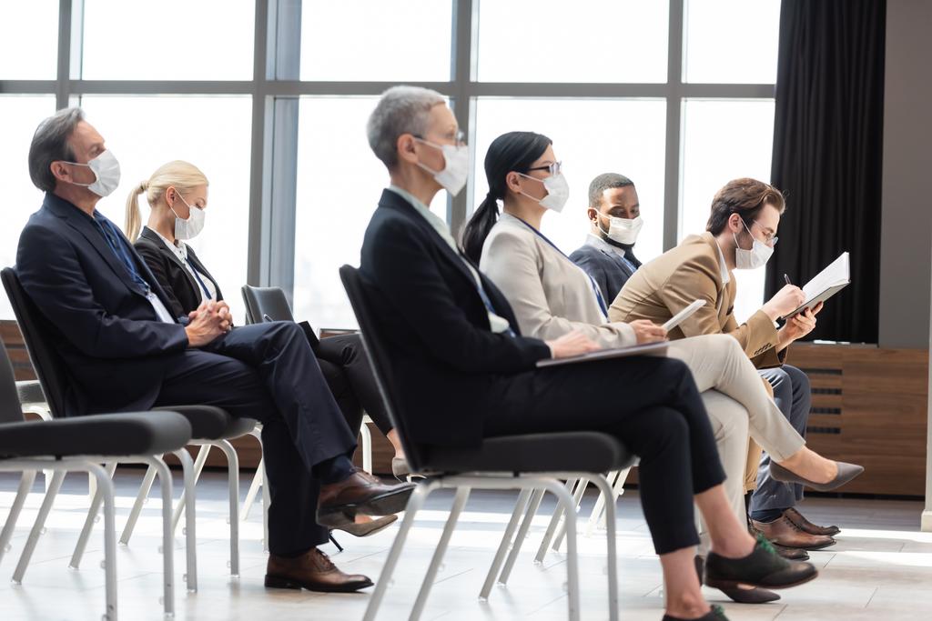 мультикультурные бизнесмены в медицинских масках сидят в конференц-зале во время семинара - Фото, изображение