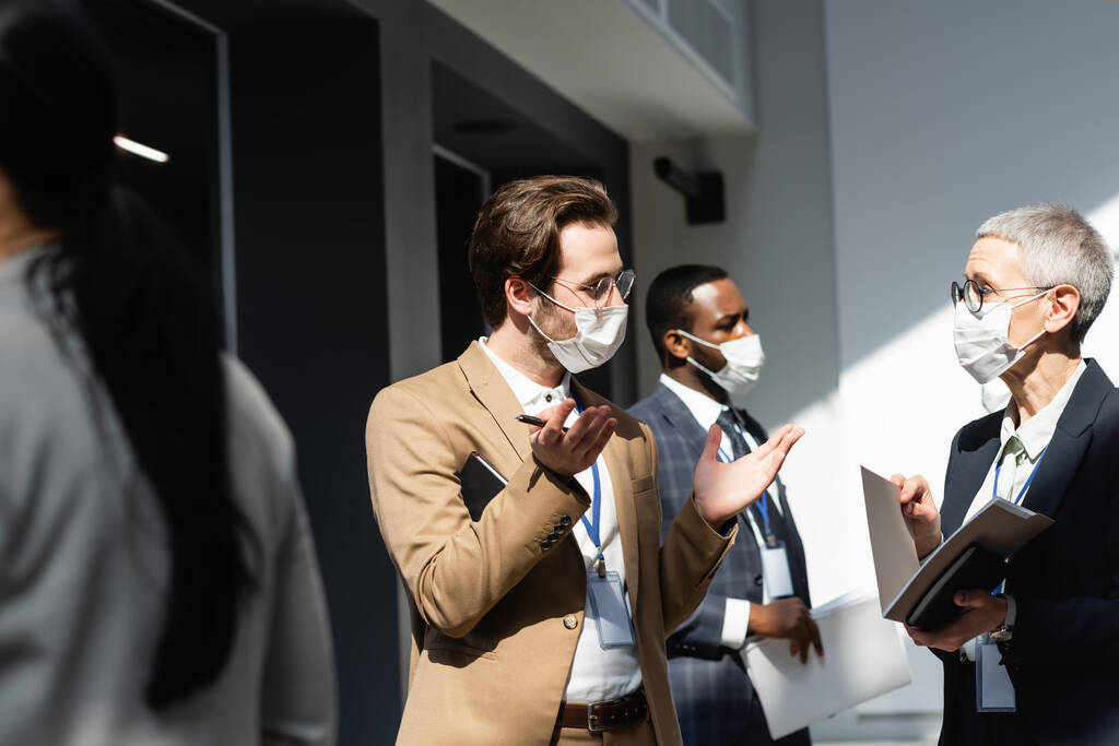 молодий бізнесмен в медичній масці жестикулює біля зрілої бізнес-леді і міжрасових колег
 - Фото, зображення