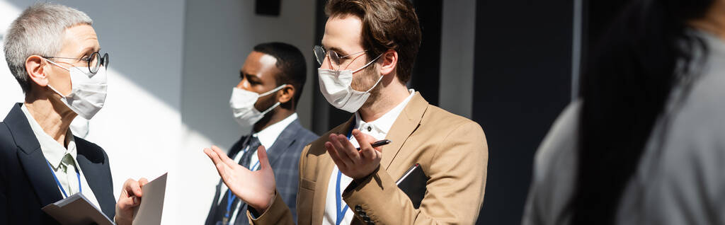 young businessman in medical mask talking to mature colleague, banner - Photo, Image
