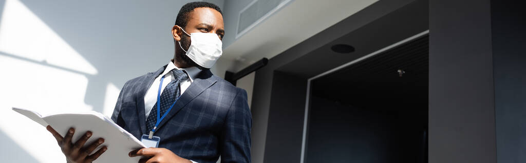 Афріканський американський бізнесмен у медичній масці тримає документи, озираючись на офіс, банер. - Фото, зображення