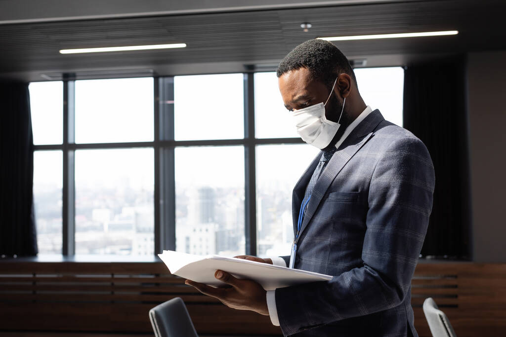 Afrikanischer amerikanischer Geschäftsmann mit Schutzmaske schaut sich Dokumente im Büro an - Foto, Bild