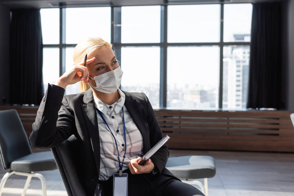 giovane donna d'affari in maschera di sicurezza guardando lontano mentre seduto in sala conferenze con notebook - Foto, immagini