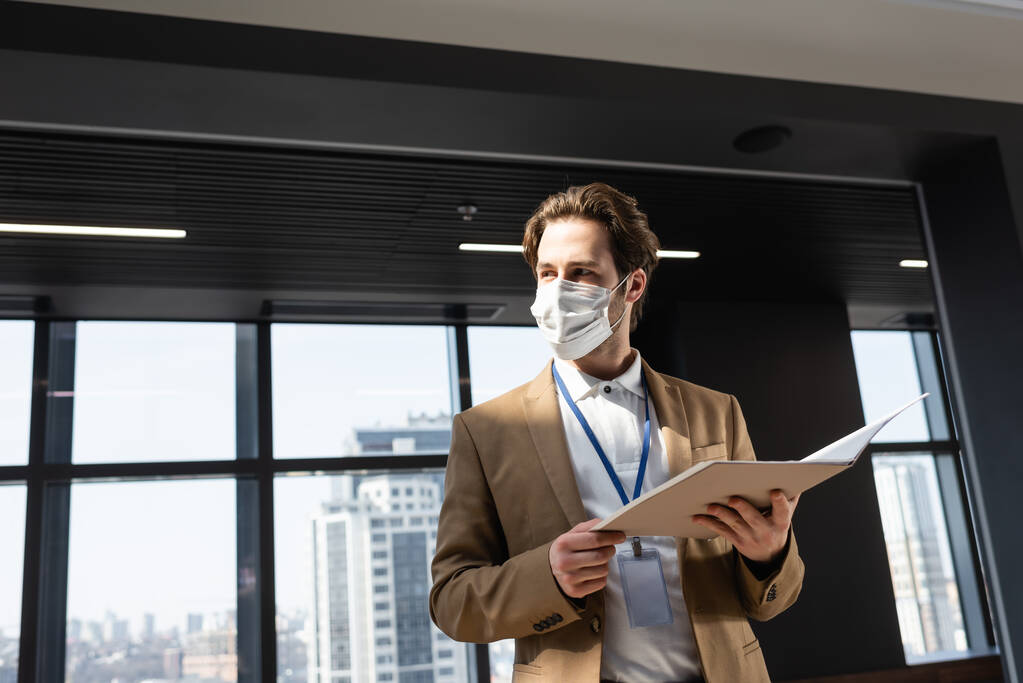 biztonsági maszkos üzletember dokumentumokkal, miközben félrenéz az irodában. - Fotó, kép