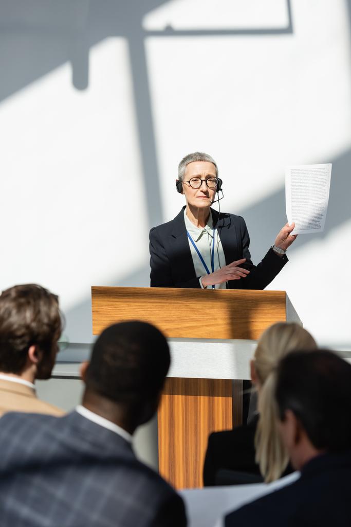 Keski-ikäinen puhuja osoittaa asiakirjaan, joka on lähellä hämärtynyttä yleisöä yrityskonferenssissa - Valokuva, kuva
