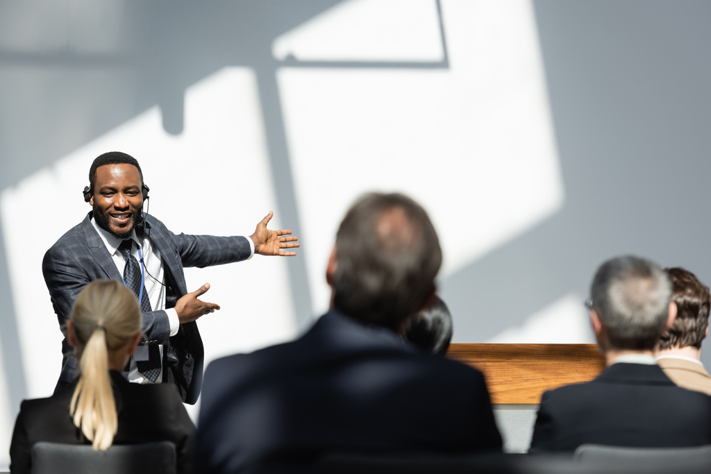 усміхнений афроамериканський викладач вказує руками біля бізнесменів на розмитому передньому плані
 - Фото, зображення