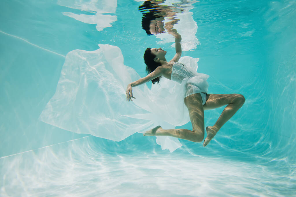 грациозная молодая женщина в платье плавает в бассейне с голубой водой  - Фото, изображение