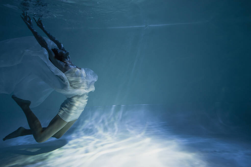 青い水でプールで泳ぐ白いエレガントなドレスの若い優雅な女性の近くの照明  - 写真・画像