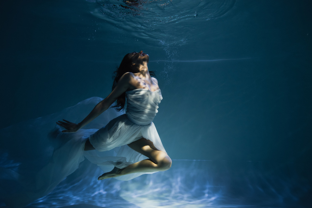 φωτισμός σε νεαρή χαριτωμένη γυναίκα σε λευκό κομψό φόρεμα κολύμπι στην πισίνα με μπλε νερό  - Φωτογραφία, εικόνα