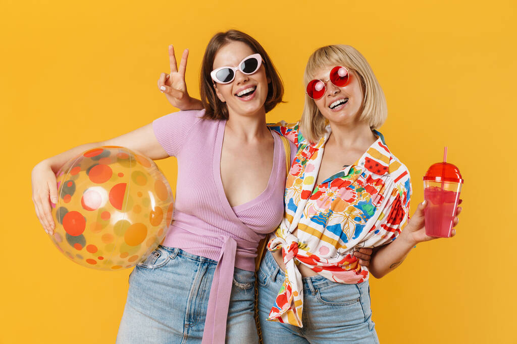 Portret van twee vrolijke lieftallige vriendinnen in zomerkleding, geïsoleerd over gele achtergrond, strandbal vasthoudend, drank drinkend - Foto, afbeelding
