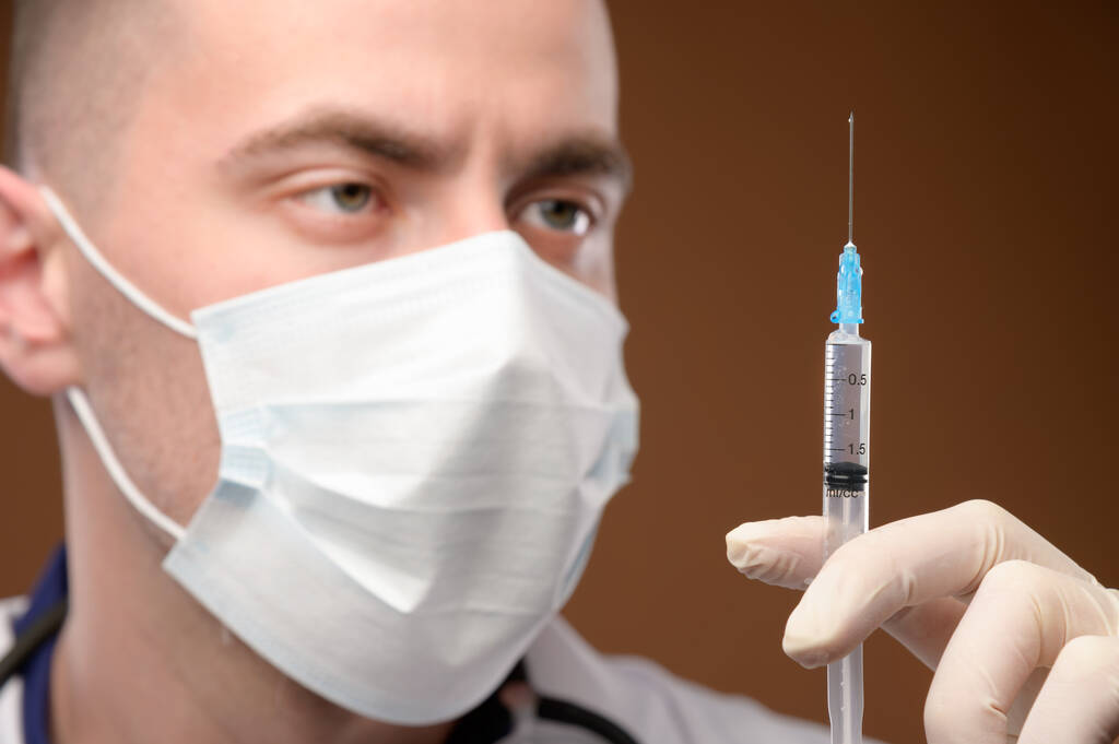 A young male Caucasian doctor in a mask holds a syringe with an injection with his gloved hand. Vaccines and vaccinations and injections concept. Focus on the syringe - Photo, Image