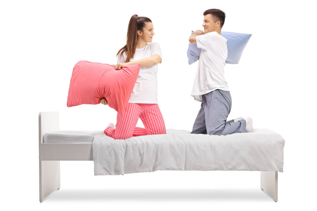 Young man and woman in pajamas fighting with pillows on a bed isolated on white background - Photo, Image