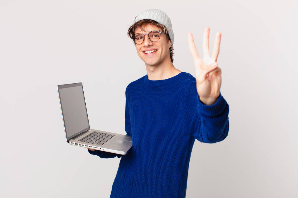 jeune homme avec un ordinateur portable souriant et regardant amical, montrant numéro trois - Photo, image
