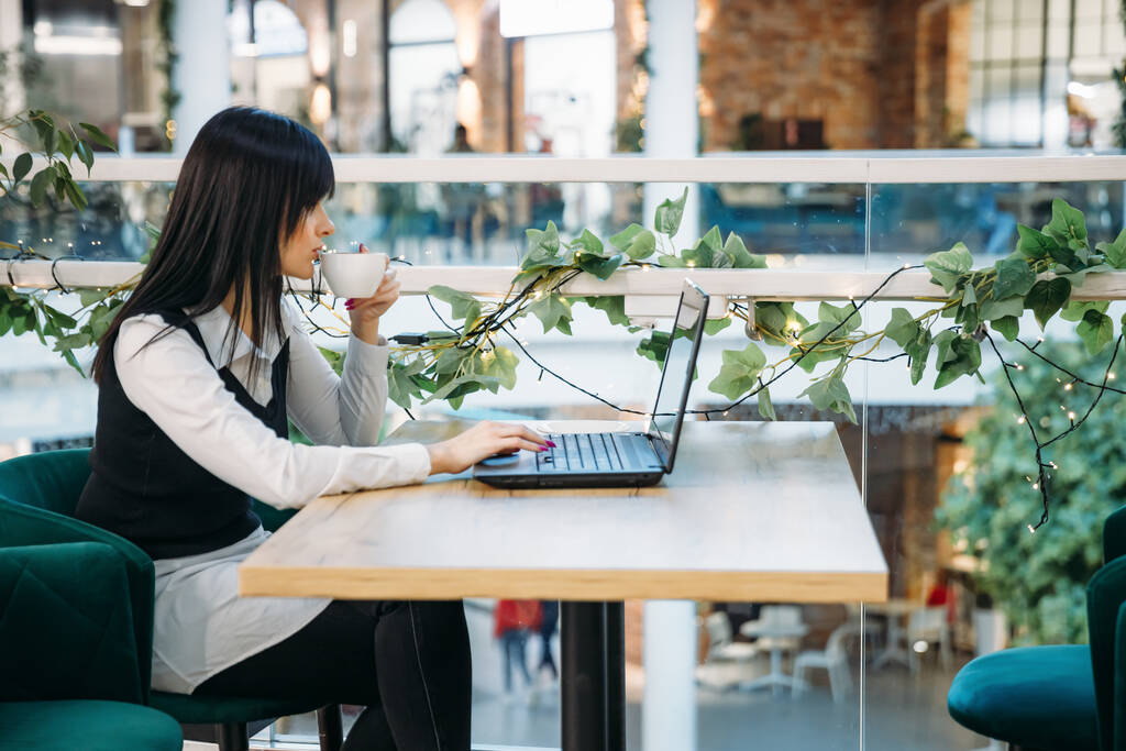 Вид сбоку молодой деловой женщины, работающей за компьютером в кафе. Пьет кофе во время работы - Фото, изображение