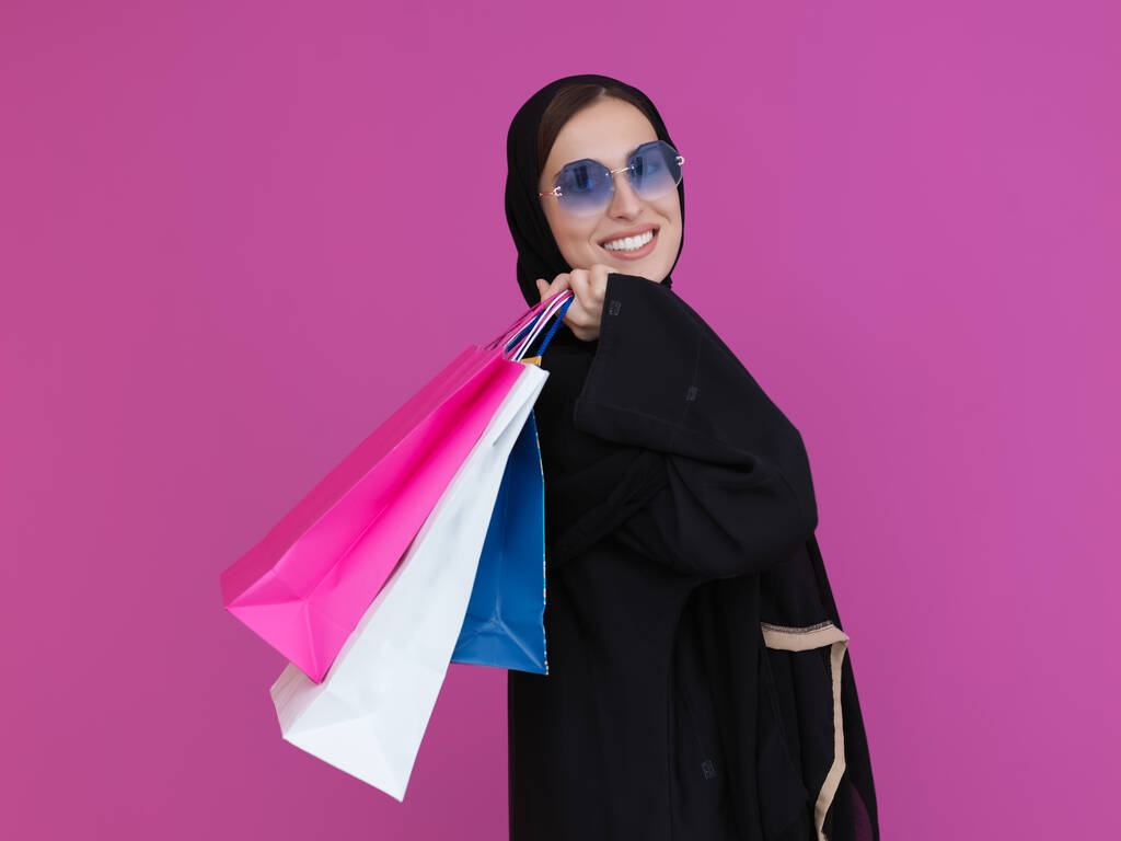 Szczęśliwa muzułmanka pozująca z torbami na zakupy. Arabska kobieta ubrana w tradycyjne czarne ubrania i okulary przeciwsłoneczne reprezentujące bogaty i luksusowy styl życia - Zdjęcie, obraz