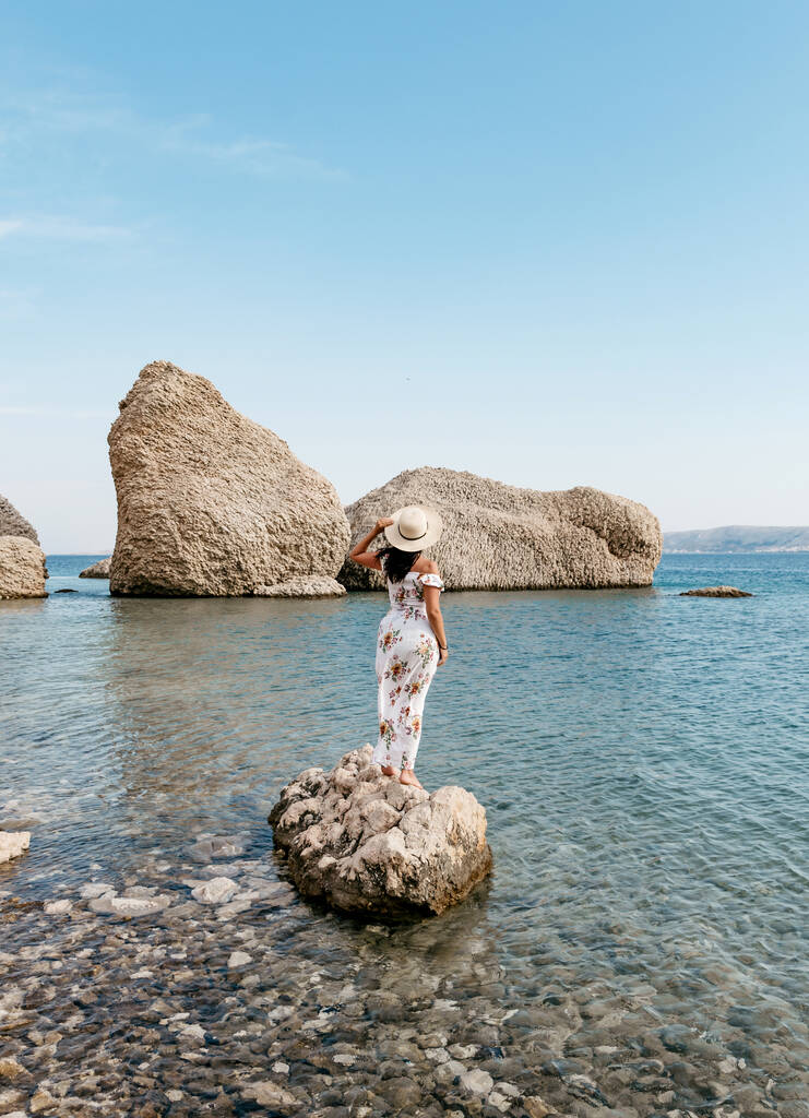 エレガントな長い白い夏のドレスの女性はクロアチアのパグ島に大きな岩の形成を持つ美しいベリトニカビーチの岩の上に立っている. - 写真・画像