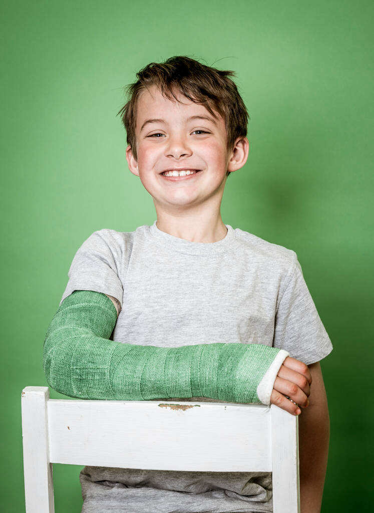 cool young schoolboy with broken arm and green plaster is sitting on white chair in front of green background - Photo, Image
