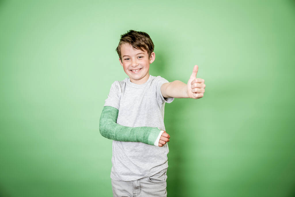 cool jovem estudante com braço quebrado e gesso braço verde posando na frente de fundo verde - Foto, Imagem
