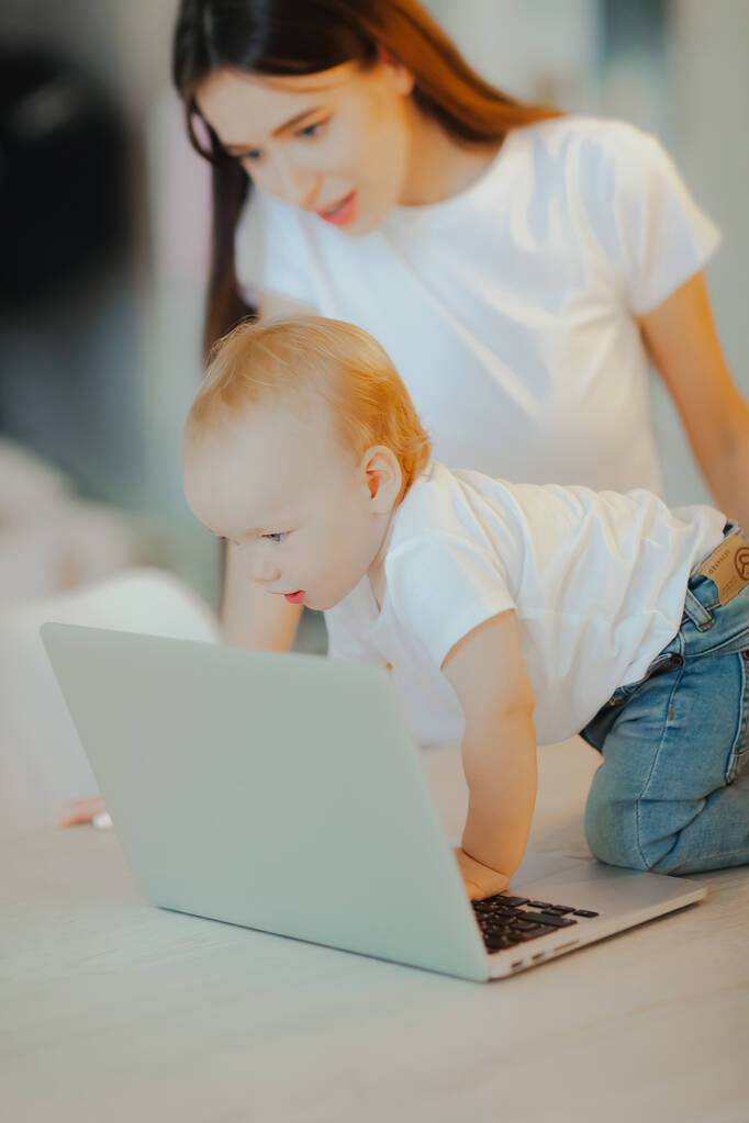 小さな子供はノートパソコンで遊ぶ一方、お母さんは彼の世話をする。ベビーケアとテクノロジー - 写真・画像