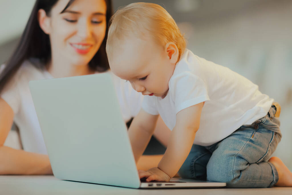小さな子供はノートパソコンで遊ぶ一方、お母さんは彼の世話をする。ベビーケアとテクノロジー - 写真・画像