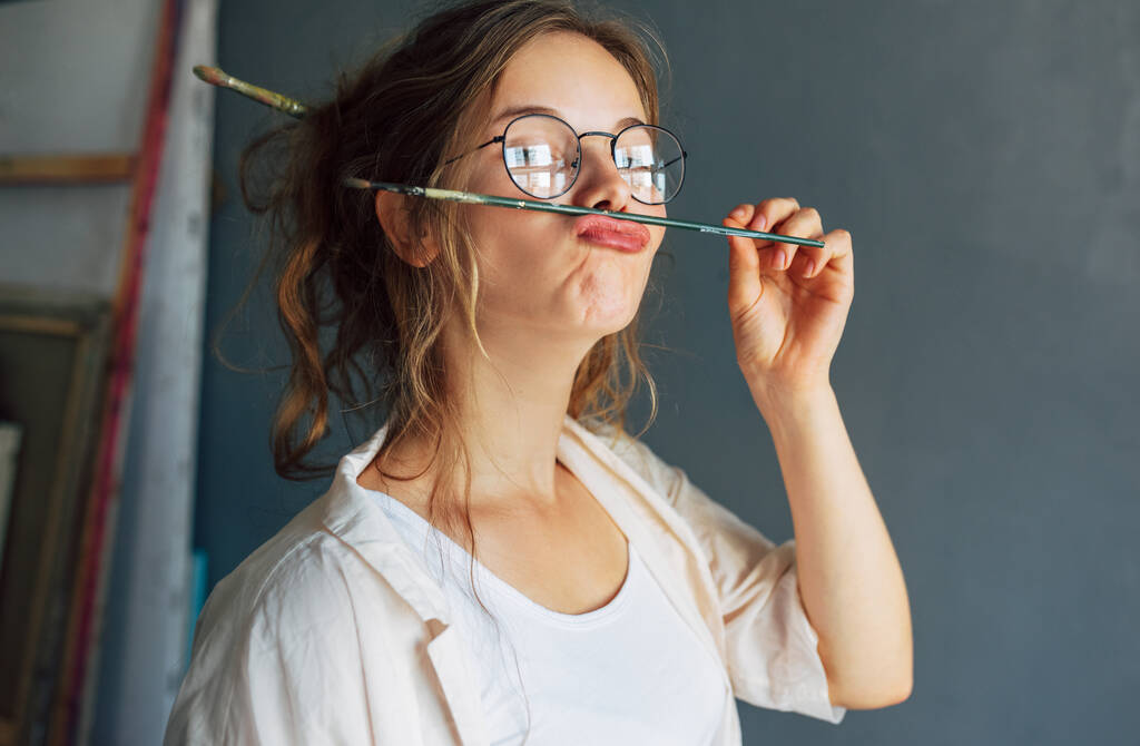 Porträt einer lustigen schönen Künstlerin mit Brille, die in ihrer Werkstatt einen Pinsel auf den Lippen hält. Hübsche junge Malerin macht Fratzen mit Pinsel in ihrem Kunstatelier. - Foto, Bild