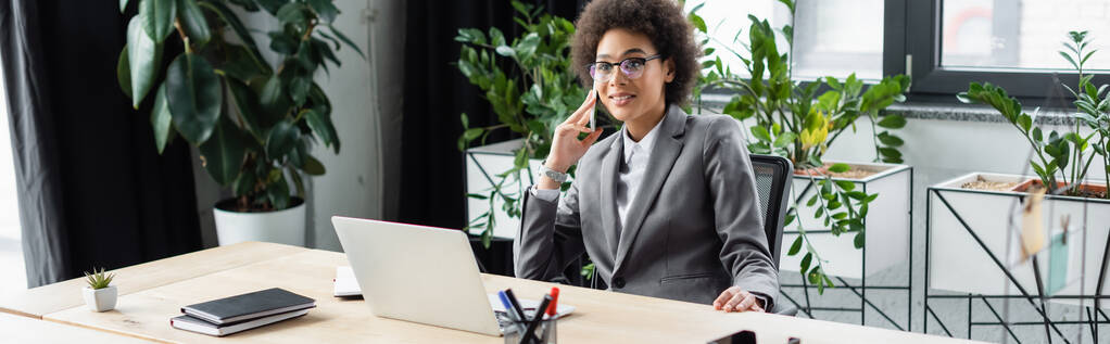 Sonriente mujer de negocios afroamericana hablando en el teléfono inteligente cerca de la computadora portátil en la mesa, pancarta  - Foto, Imagen