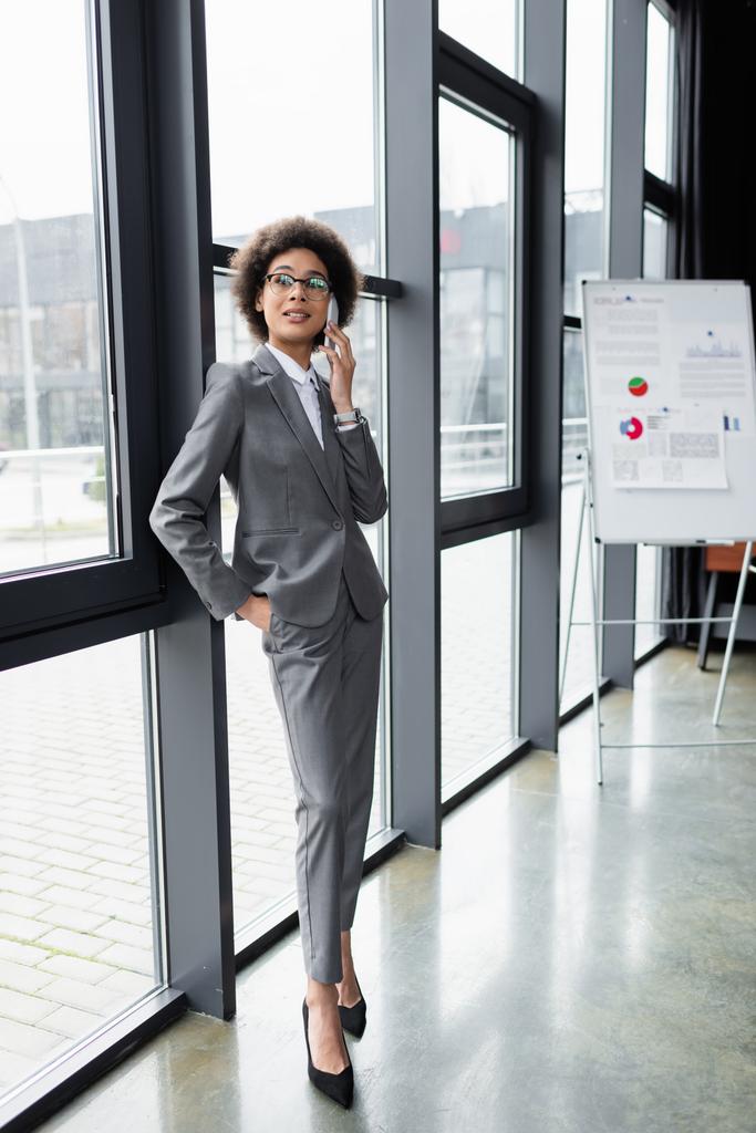 Empresaria afroamericana positiva hablando por celular cerca de la ventana en la oficina  - Foto, Imagen