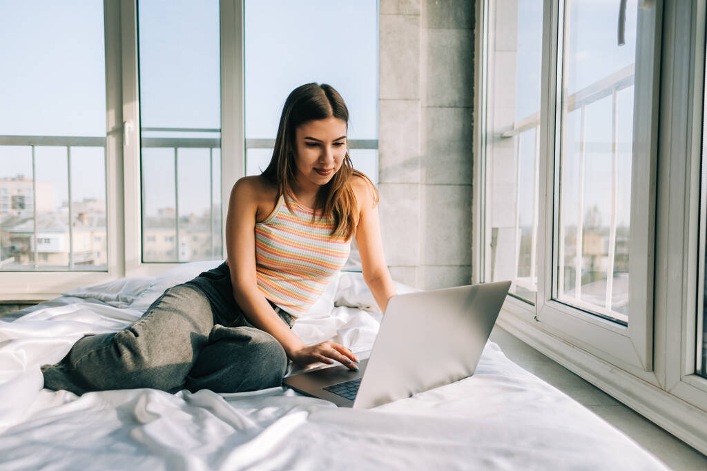 Young caucasian woman resting on a bed at home and using laptop computer. Chatting with friends, shopping online.	 - Foto, Bild