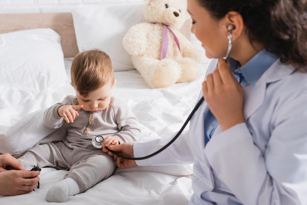 pédiatre afro-américain flou en manteau blanc examinant bébé garçon avec stéthoscope près de la mère - Photo, image