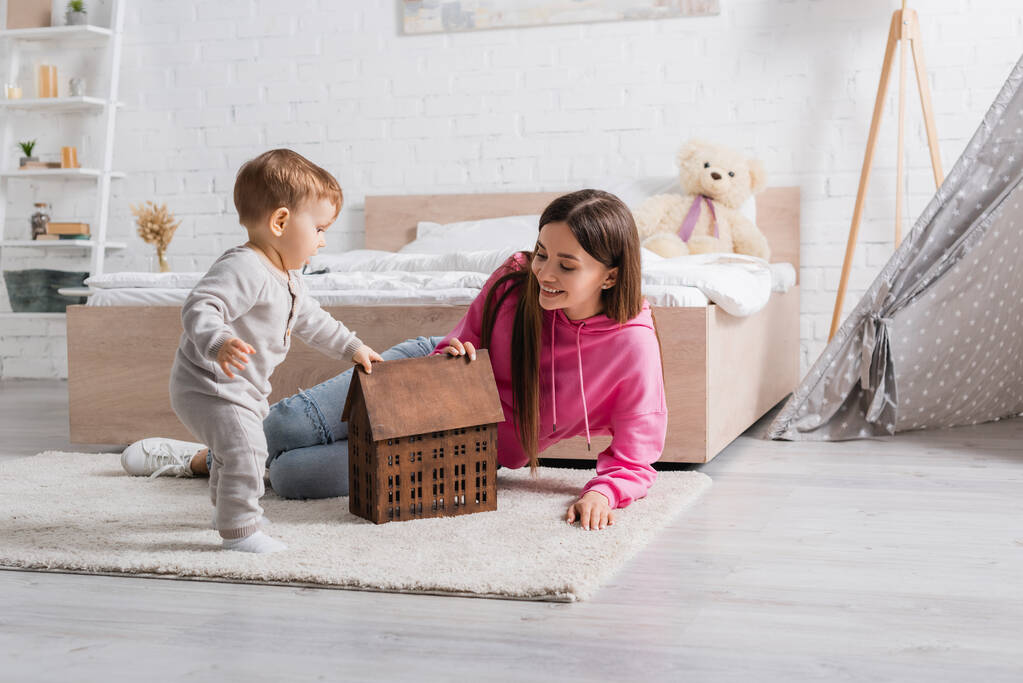 счастливая мать смотрит на модель дома рядом с младенцем сыном на ковре  - Фото, изображение