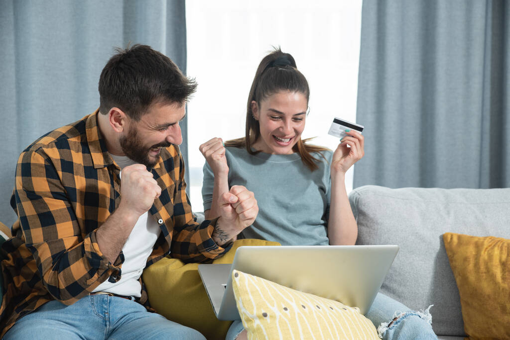 Das junge glückliche Paar sitzt in seiner Wohnung und ist glücklich, weil es es nach langer Zeit der Selbstisolierung und Quarantäne geschafft hat, Reisen und Urlaub auf seinem Laptop über das Internet zu buchen. - Foto, Bild