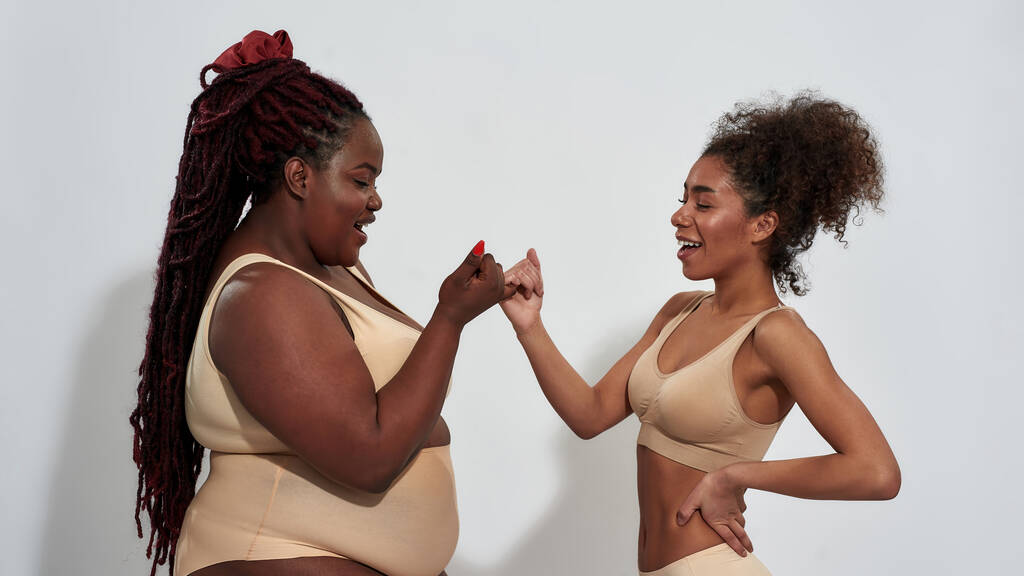 Дві радісні афро-американські жінки в білизні з різним розміром тіла сміються, роблячи рожеву обіцянку, стоячи ізольовано над сірим фоном. - Фото, зображення