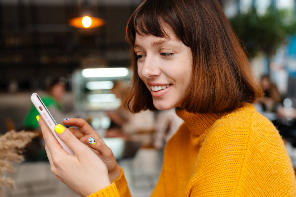 Menina ruiva feliz sorrindo e usando o celular enquanto sentado no café dentro de casa - Foto, Imagem