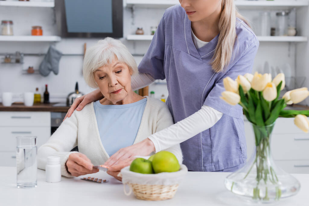 trabajadora social señalando píldoras en manos de una mujer mayor sentada en la cocina - Foto, imagen