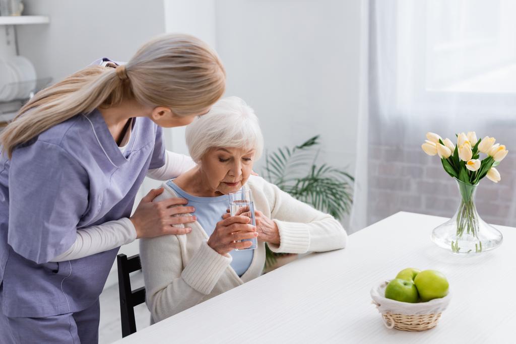 νεαρή νοσοκόμα αγκαλιάζει τους ώμους της ηλικιωμένης γυναίκας κρατώντας ένα ποτήρι νερό, ενώ κάθεται κοντά στο τραπέζι - Φωτογραφία, εικόνα