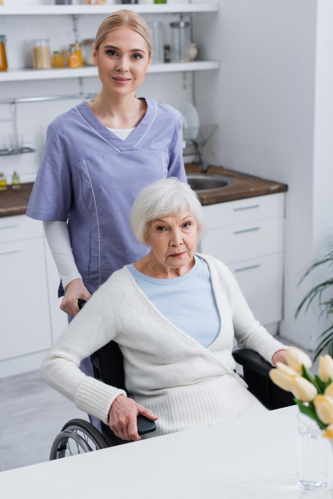 молодая медсестра и пожилая женщина-инвалид смотрит в камеру на кухне - Фото, изображение